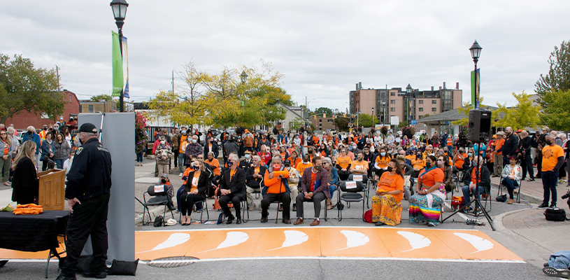 A crowd of people gather in front of a crosswalk painted orange with 7 white feathers.